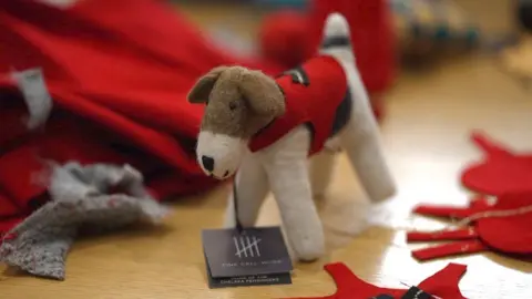 A little white and brown felt dog wearing a red jacket stands on a table among offcuts of other bits of fabric