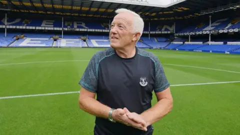 Everton FC Bob Lennon is standing pitchside at Goodison Park. He looking off camera and is wearing an Everton T-shirt.