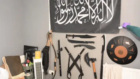 West Midlands Police A wall in a bedroom covered in weapons including swords, an axe and a bow. A black banner has been hung above them.