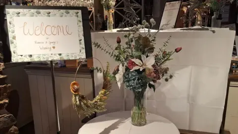 Penny Farthing Welcome sign at entrance to restaurant with the couple's names and a round table with white table cloth and bunch of flowers in a glass vase. A wreath made of flowers and grasses hangs from the welcome sign 