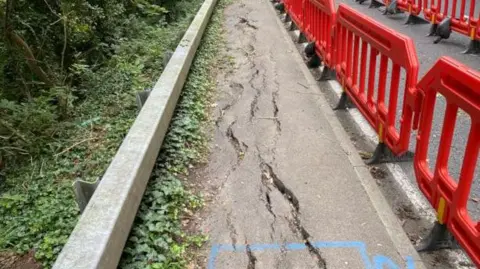 National Highways Cracking in the footpath can be seen alongside the A36. The path is uneven. Red traffic bollard can be seen between the bath and the carriageway.