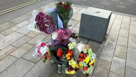 Suzy Lawson / BBC Bouquets of flowers in a range of colours placed on a pavement next to a square concrete block. There is a silver can of Coors lager also there.