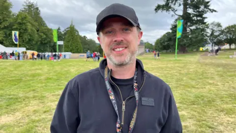 Dougie Brown is standing outdoors at the Belladrum festival site near Beauly. He has a trimmed beard and is wearing a black cap and back fleece over a black T-shirt.