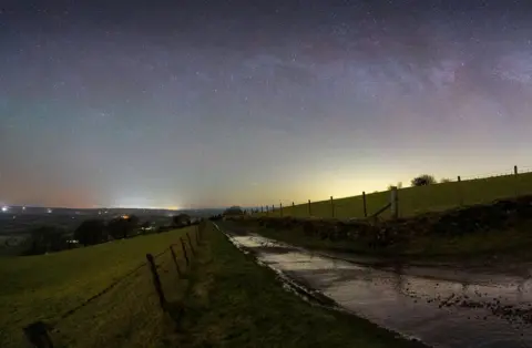 Serydda/Dafydd Wyn Morgan The Milky Way shines above a field and wooden fence. Light pollution is visible on the horizon. 