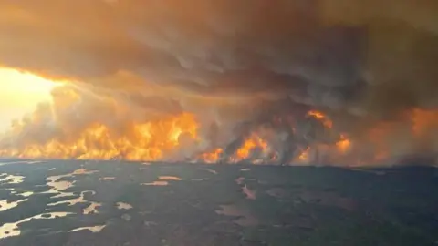 Image taken from a plane. You can see a wildfire on the horizon, with flames and smoke covering the landscape