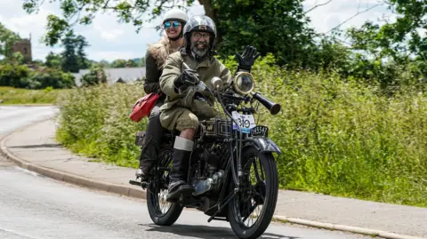 Peter Wileman A man and woman on a vintage motorbike. The man has a beard and black glasses and is waving to the camera. The woman, with blonde hair sat behind him is wearing sunglasses and smiling towards the camera. both are wearing helmets