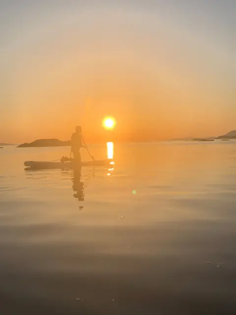 Kirsten Lyons A paddle boarder goes across water with the sun setting, creating an orange hue in the sky