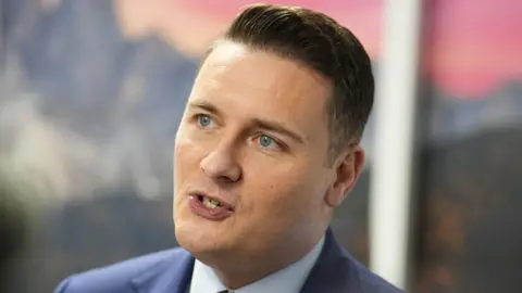 Health Secretary Wes Streeting talking during a media interview. He has short dark hair and wears a navy suit with a white shirt and red tie underneath.