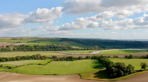 a countryside landscape, showing green fields bordered by trees and hedges