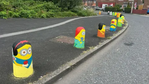 Brightly-coloured Minions have been painted on to bollards on a pavement. Minions are small, yellow, capsule-shaped fictional creatures from the Despicable Me films.