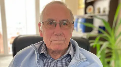 Family handout Richard Lee looks into the camera while seated in his lounge. He has receding, short and neat grey hair and metal rimmed glasses. He wears a pale blue zipped fleece over a check, buttoned shirt.