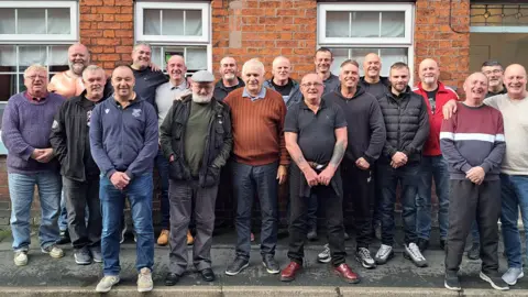 LEEK CALENDAR BOYS 2025 Twenty men stand on the pavement, gathering in a huddle outside a brick building. Three window panes and a door can be seen behind them. They are all dressed differently, mainly in jackets, dark jeans and trainers.