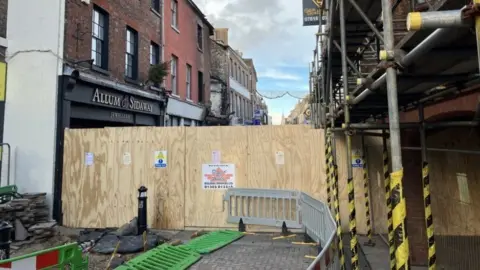 Wooden hoarding cloking off street with buildings both sides and scaffolding covering a building on the right.