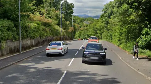 Google Three cars drive along Clune Brae while a woman walks on the pavement