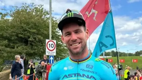Sir Mark Cavendish in his blue 35 stage victory jersey at the sports centre raceway. A Manx flag is behind him with crowds.