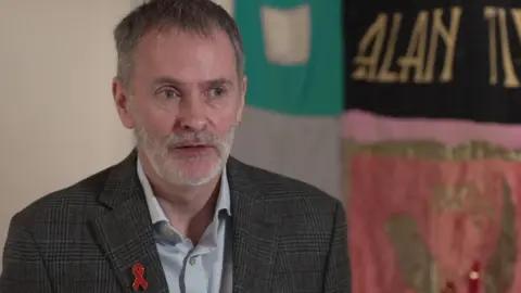 Andrew Given speaking on camera inside the museum, wearing a jacket with a red ribbon pin.