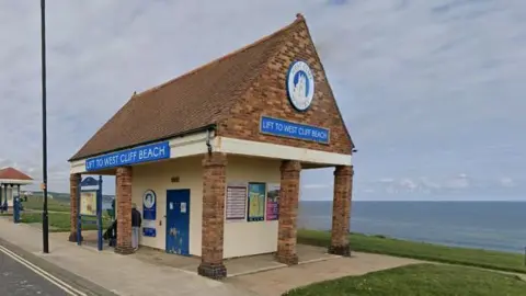 Google Whitby Cliff Lift - a red-brick building with a pointed roof and blue signage reading 'Lift to West Cliff Beach'. The building is overlooking a view of the sea.