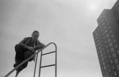 Sandra George Estate A boy in dark trousers and a light jacket sits on a climbing frame in front of the high flats of Greendykes. The sun is seen above the building. 