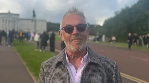 Paul corry is pictured in the Stormont Estate. He has short white hair and a beard and is facing the camera wearing sunglasses. Behind him is the Stormont building in the distance and a large number of people, all are out of focus.