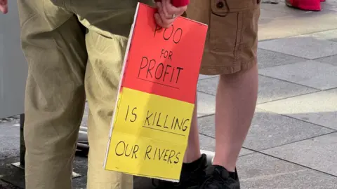 A yellow and red sign saying Poo For Profit - Is Killing Our Rivers is being held by a man in light brown trousers