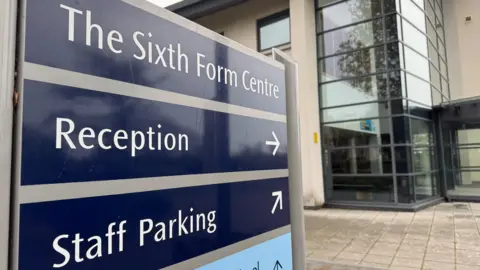 A sign at a sixth form in Guernsey. There is a building in the background made from glass and stone.