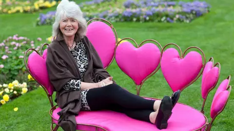Getty Images Dame Jilly Cooper on a heart-shaped pink sofa with her feet up, in dark clothes and a leopard-skin top