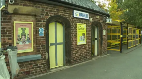 A red brick station building has a number of posters and a sign saying 'Welcome to Maghull'. There is a covered waiting area further down the platform.