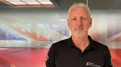 Head and shoulders shot of a man with grey hair and goatee beard standing in front of a Union flag. He is wearing a black polo shirt and has his arms behind his back.