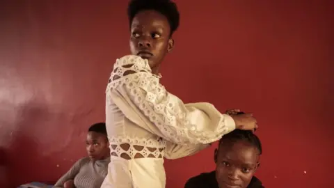 AFP A woman doing another woman's hair backstage at the Miss Freedom Day fashion show. One woman is standing up and wearing white. She is looking into the distance as she does someone's hair, who is sitting down. There is a red wall behind them.