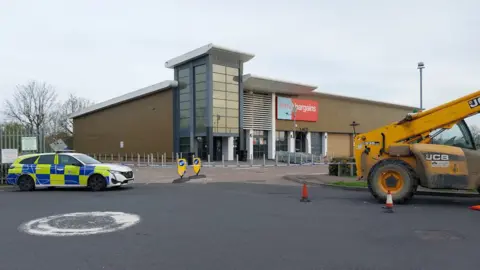 A discount store with a police cordon around it and a police car parked on the left and a yellow digger on the left.