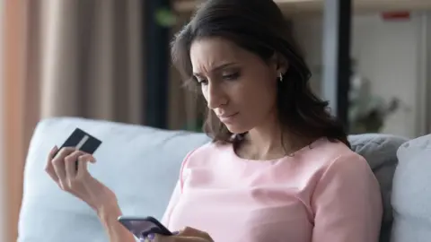 Getty Images A concerned woman checks her phone while holding a debit card