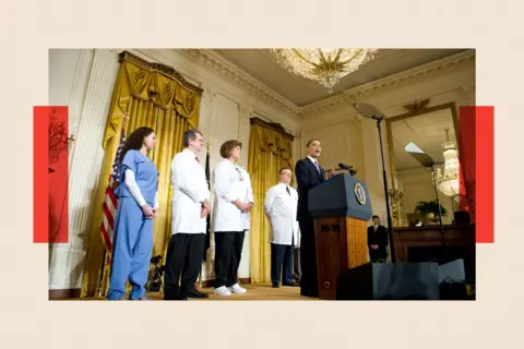 Getty Images Three people in white medical coats, and one woman wearing blue hospital scrubs, stand behind Barack Obama on a stage in the White House East Room as he makes a policy announcement