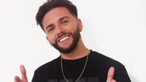 Leon Mallett posing with his fingers pointing towards the camera. He has black hair, a short beard and is wearing a black t-shirt and silver necklace 