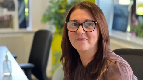 Charity managing director Sue Brennan sits in an office chair and smiles at the camera, she wears a brown satin striped blouse and has long dark hair and glasses.