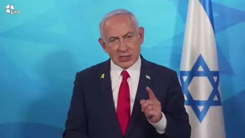 Kantor pers pemerintah Israel Benjamin Netanyahu melihat lurus ke bawah kamera, menunjuk dengan satu jari. Bendera Israel menggantung di belakangnya.