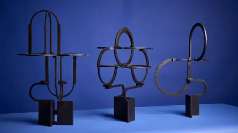 New Designers/Mark Cocksedge Three of Mr Pearson's sculptures pictured on a blue table. The sculptures are made from metal and are mounted on plinths.