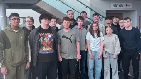 A group of 15 students, mostly wearing hoodies, jeans, t-shirts and jogging bottoms are lined up and looking directly at the camera in front of a flight of glass and metal stairs in a foyer.