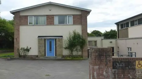 St Bernadette's Parish House, on Bowerham Road. A large detached house with a blue front door and two windows on the second floor. The house is rendered and red brick and has a large asphalt drive to the front.