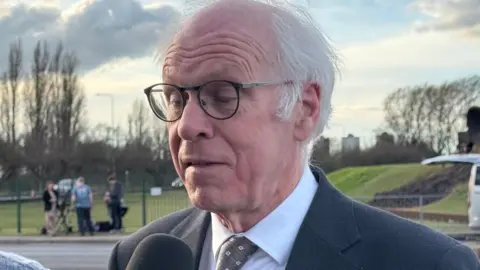 BBC/Dale Baxter Nic Dakin in a dark suit and patterned tie stands outdoors by a roadside, speaking into a handheld microphone. A van, grassy embankment, and distant figures are visible in the background under a cloudy sky, suggesting a media interview taking place.