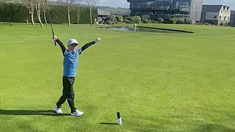 Official Hole in 1 Freddie wearing a light grey cap and blue gold top and black golf pants and white golf shoes throws his arms up in the air to celebrate his hole-in-one on Carus Green Golf Club course.