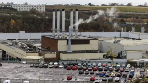 Getty Images A drone shot shows the scale of the Vauxhall factory in Luton. In the centre is the plant itself with smoke coming from a large flue on the roof. There is a busy car park in the foreground and the runway of Luton Airport, with a plan on it, can be seen on the horizon.