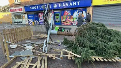 A Christmas tree is collapsed on the pavement in a shopping High Street. There is a wooden fence around the tree which has been demolished in places and the tree is wilted on its side. There are convenience shops in the background behind the tree.