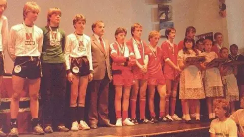 The Orkney U16s football team standing on stage with their bronze medals in 1985, the first year of the Island Games.