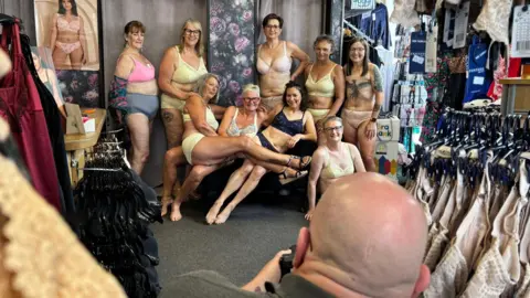 Taunton Breast Friends A group of women stand, sit and lay down in a shop, all wearing their underwear. A man's head is seen in the foreground, and he can be seen holding a camera.