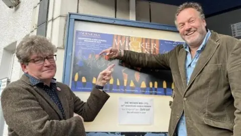 Two men in their 50s either side of a poster for The Wicker Man outside a cinema. Dominic, on the left, is in a tweed suit and has glasses and thick hair. Justin (right) is in a cord suit and has a beard. They are smiling and pointing at the poster.