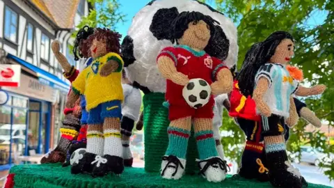 Beryl's Purls Knitted and crocheted mini footballers in colourful kits from countries including Argentina, Portugal, Brazil and Spain stand on a green knitted base on a flat surface. Rows of shops can be seen behind.