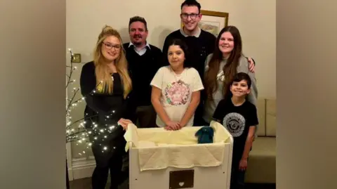 Chelsea Turrell, Christian Horn, and their children Aria, and Leo. Russell Fox, Funeral Director for the Co-operative Funeralcare Kendal, and Jennifer Plews, Funeral Arranger for the Co-operative Funeralcare Kendal. They are all standing around a cot and smiling.
