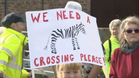 BBC Girl carrying a sign saying 'we need a crossing please'