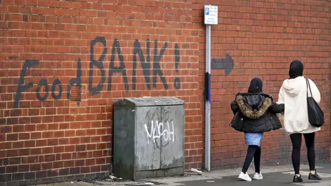 Woman and a child walking past a wall with 'food bank' painted on