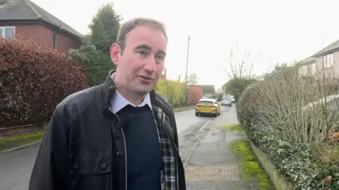 Councillor James Pearson is standing in a residential street, with parked cars, hedges and a number of houses behind him. He is wearing a wax jacket and blue jumper and white shirt. 